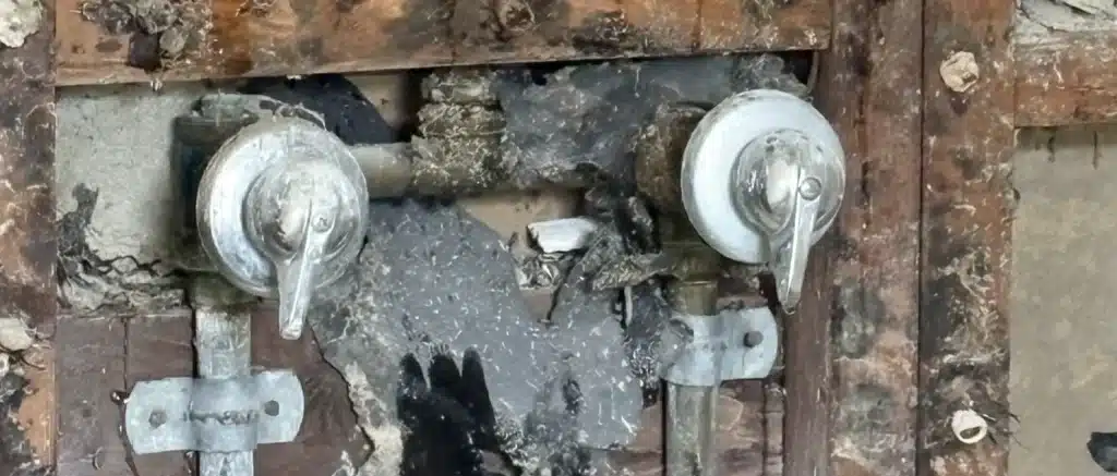 Exposed shower wall showing water-damaged framing and plumbing behind two shower controls after a hidden leak.